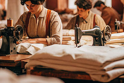 Women working at sewing machines in a historical textile workshop, stitching fabric with stacks of cloth around them.