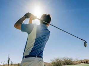 Man swinging golf club under bright blue sky.