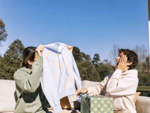 Two friends exchanging pastel hoodie gifts on sunny patio.