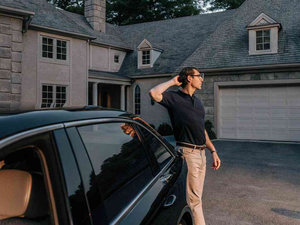 Man in black polo shirt and khaki pants standing near a black car at sunset.