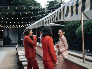 Women in red suits chatting outdoors under string lights at party.