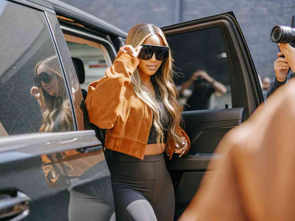 Woman in brown jacket posing beside car for paparazzi