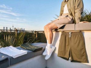 Person sitting on rooftop ledge wearing neutral shorts and jacket, surrounded by folded casual shorts.
