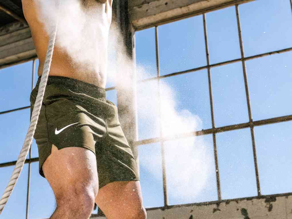 Athlete in olive green Nike shorts climbing a rope indoors with chalk dust in the air.