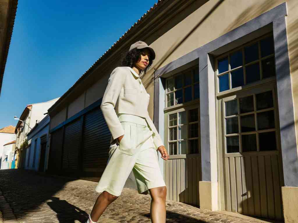 Fashionable woman in light outfit and cap walking down a sunny cobblestone street.