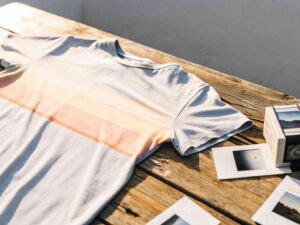 Light-colored T-shirt with pastel stripes on a wooden table next to instant photos and a retro camera.