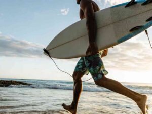 Man running into the ocean with a surfboard, wearing tropical print board shorts.