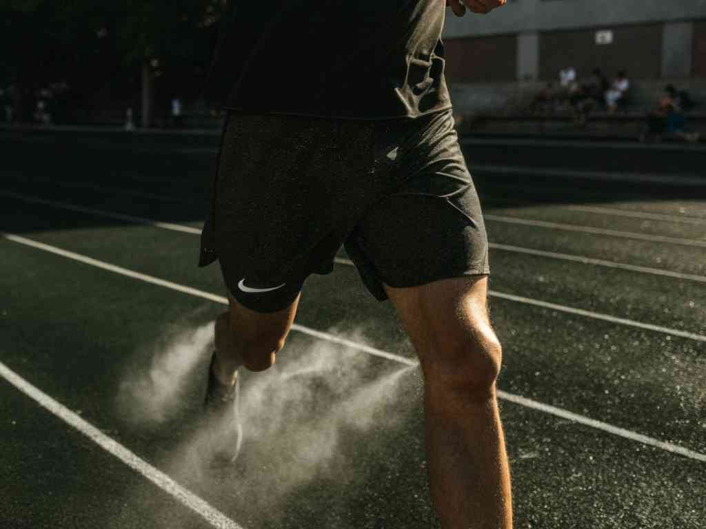 Runner wearing black Nike shorts sprinting on a track, kicking up dust in a powerful motion shot.