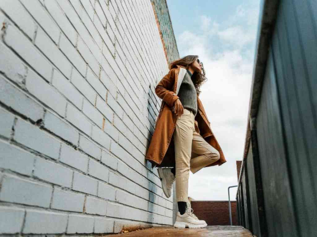 Woman in brown coat and beige pants leaning on white brick wall.