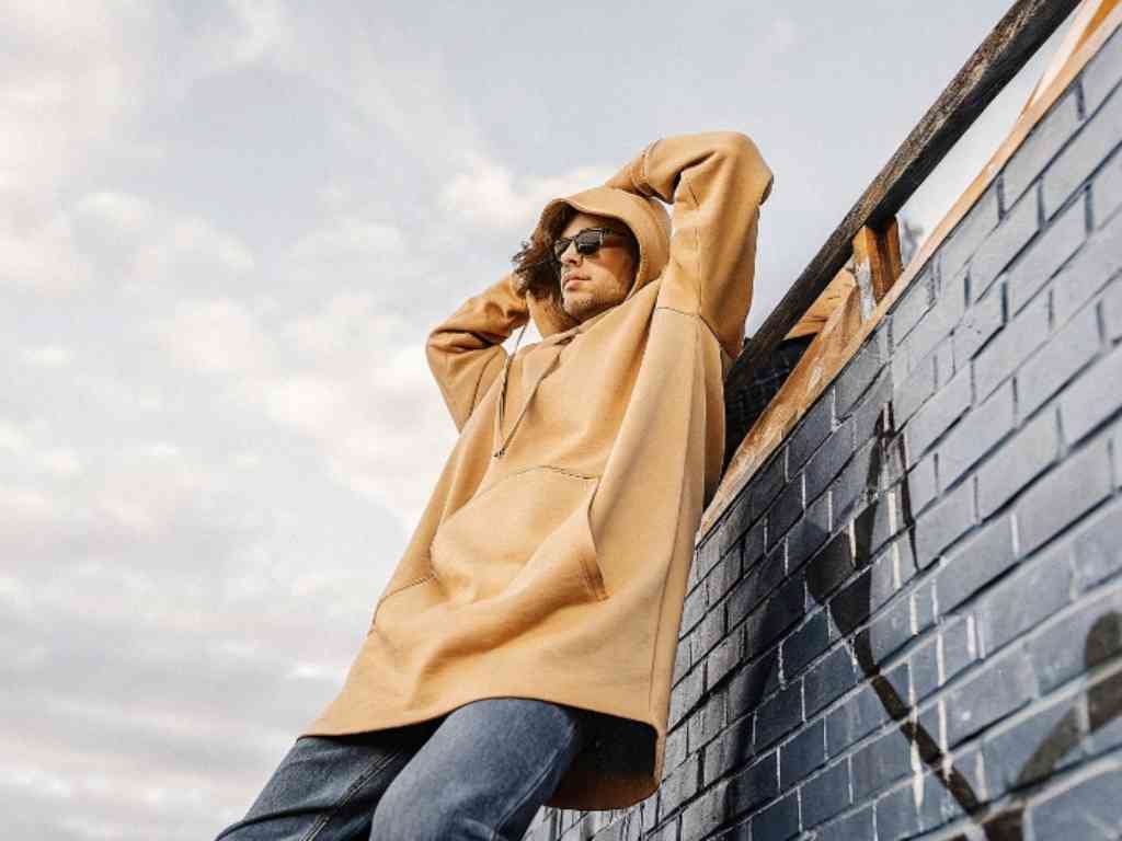 Man in oversized tan hoodie leaning against graffiti wall outdoors.