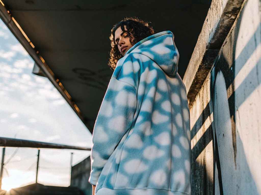 Model in oversized blue tie-dye hoodie standing in sunlight near graffiti wall.