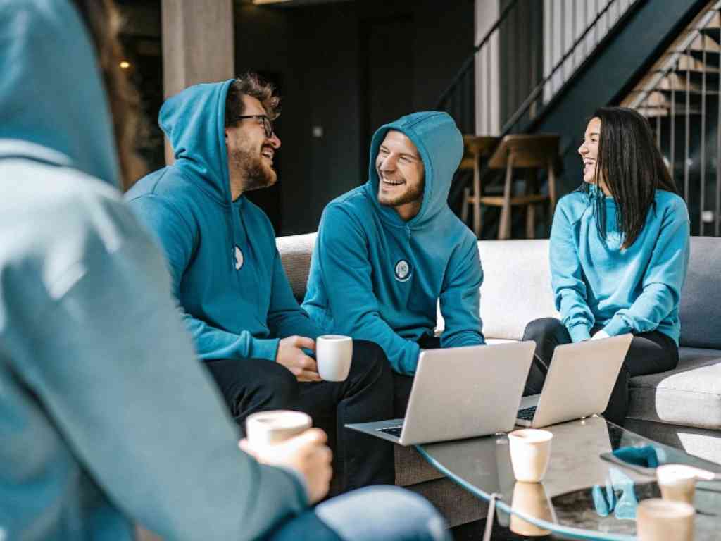 Team in blue hoodies chatting around laptops in casual lounge