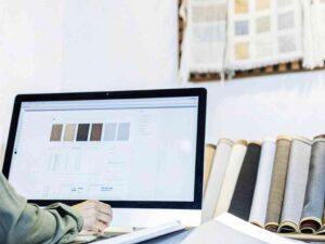Designer reviewing digital fabric samples on a desktop computer with fabric rolls in the background.