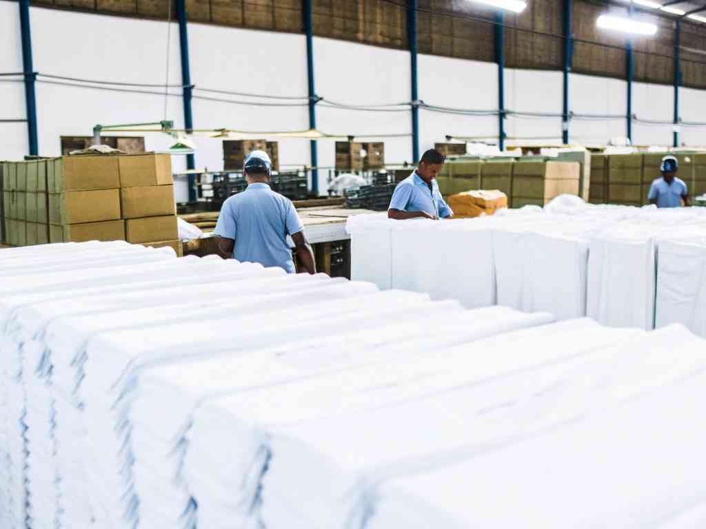 Workers in a garment factory handling large stacks of folded white fabric.
