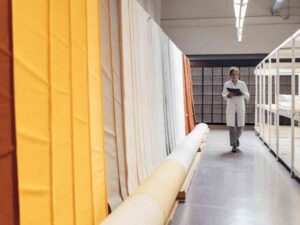 Woman in lab coat inspects colorful fabric rolls in warehouse.