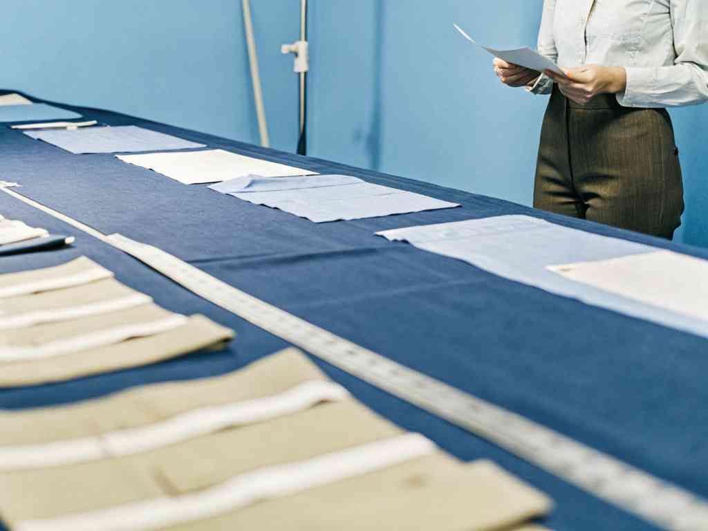 Fashion professional reviewing fabric samples and measurements on a blue table in a planning room.
