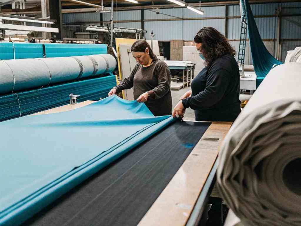 Two women handling large rolls of blue fabric in a textile production facility.