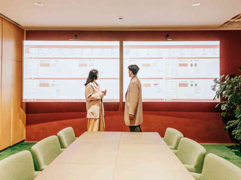 Two professionals in beige coats discussing data projected on large screens in a modern boardroom.