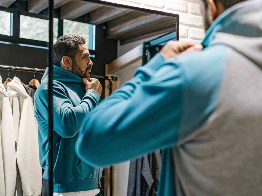 Man adjusting his hoodie in a fitting room mirror surrounded by hoodies.