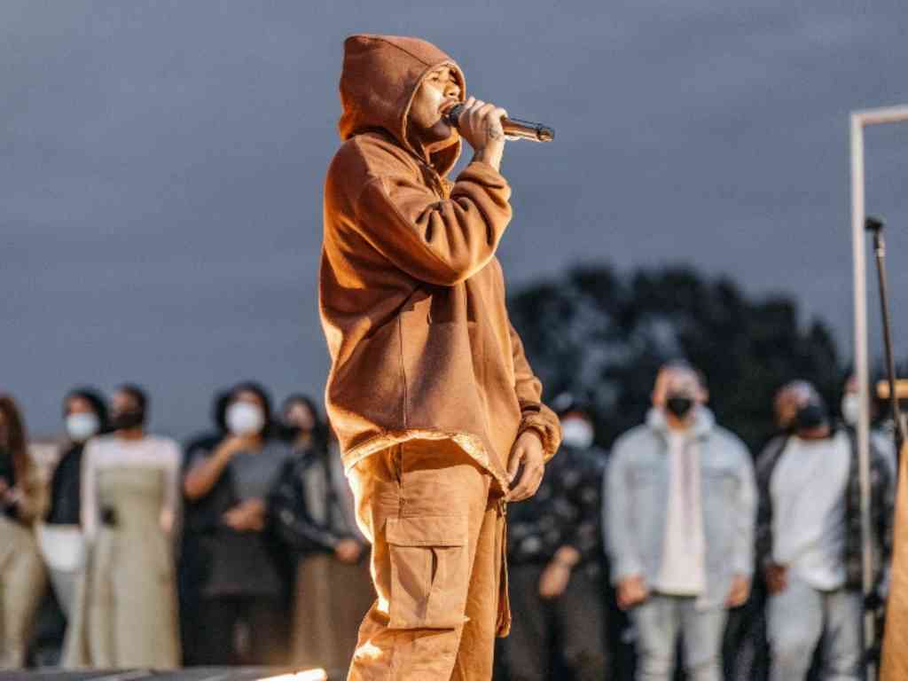 Music artist in brown oversized hoodie performs live on outdoor stage.