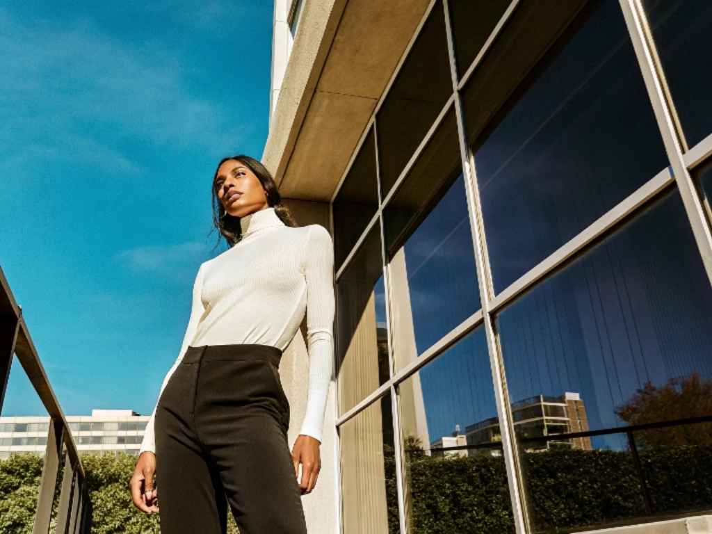 Woman in white ribbed turtleneck and black pants standing by modern glass building