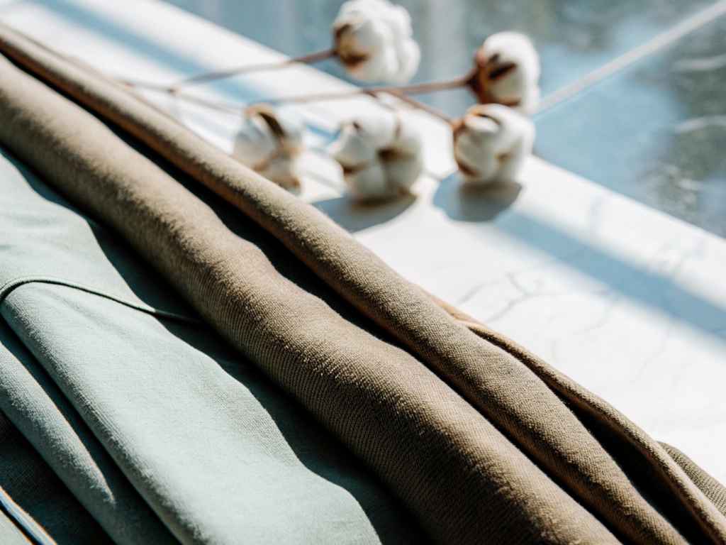 Rolled green and tan cotton fabrics beside raw cotton pods in sunlight.