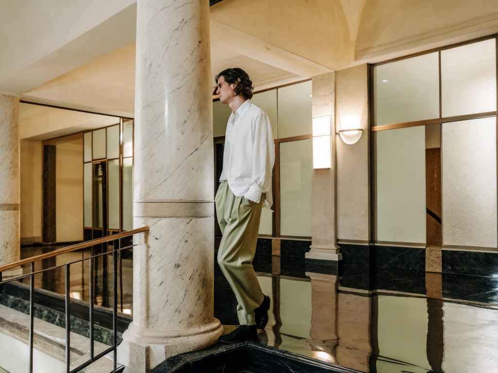 Man in white shirt and sage trousers in marble hallway.
