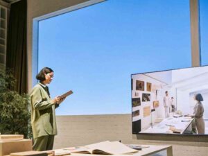 Woman in green suit presenting remotely with a tablet, large screen shows virtual design meeting.