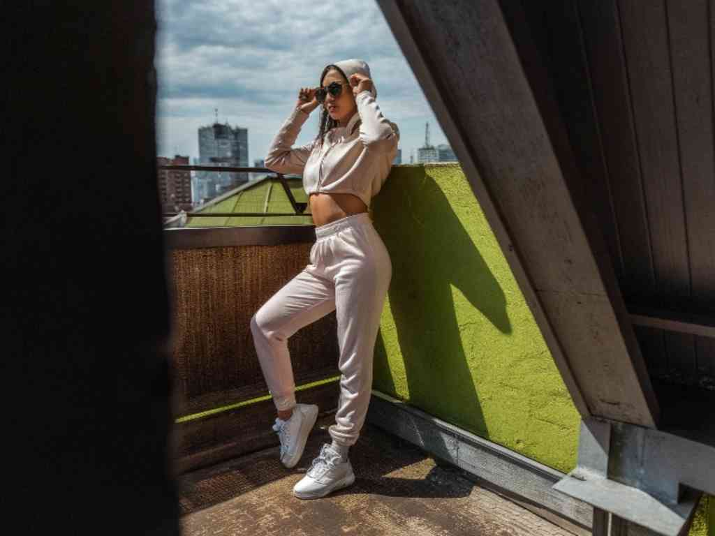 Woman in cropped hoodie and joggers posing on urban balcony in sunlight.