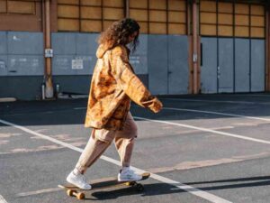Model in tie-dye hoodie skateboarding in empty parking lot.