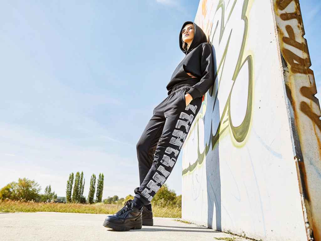 Woman in black hoodie and graphic joggers leaning on graffiti wall