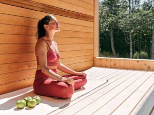 Woman in red activewear meditating on sunny wooden deck near forest.