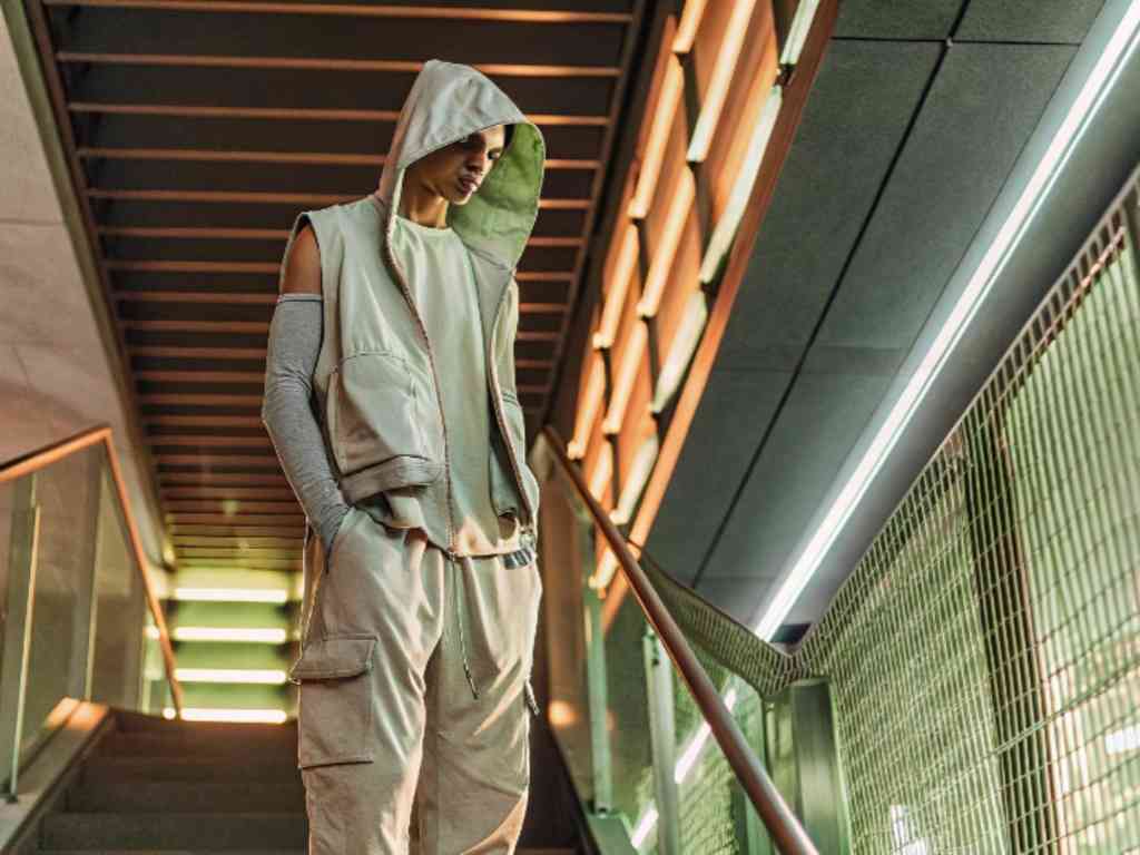 Male model in sleeveless hoodie and cargo pants walking in neon-lit corridor.