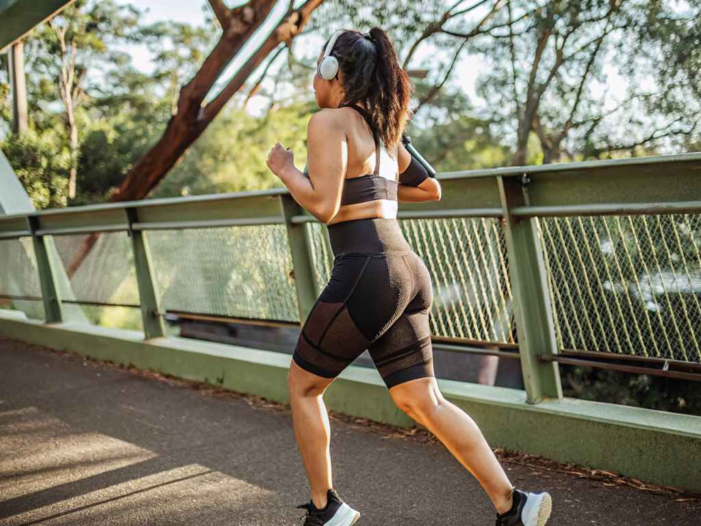 Woman in black workout set jogging across urban green bridge.