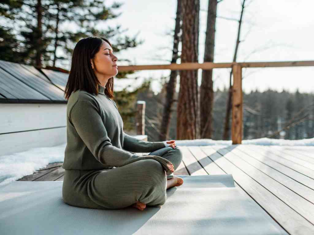 Woman in green loungewear meditating on snowy deck with forest view.