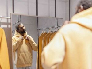 Man in a yellow hoodie adjusting the collar in front of a mirror with more hoodies behind.
