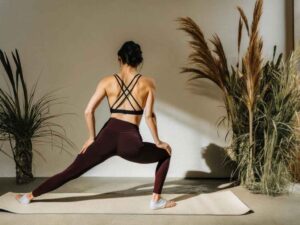 Woman in burgundy leggings and strappy sports bra stretching in sunlit yoga studio