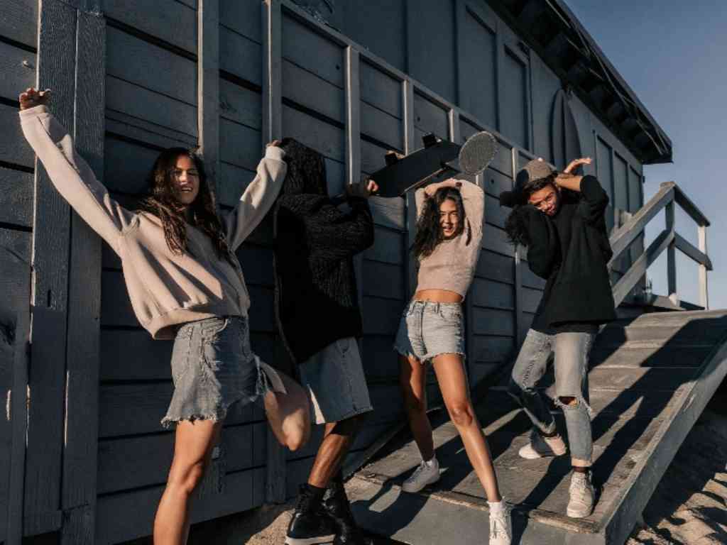 Group of young people in casual outfits dancing near a wooden beach cabin.