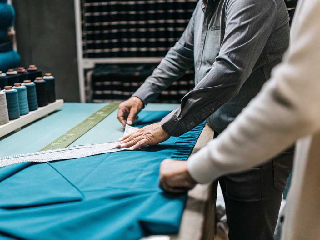 Hands measuring teal fabric beside rows of matching thread spools