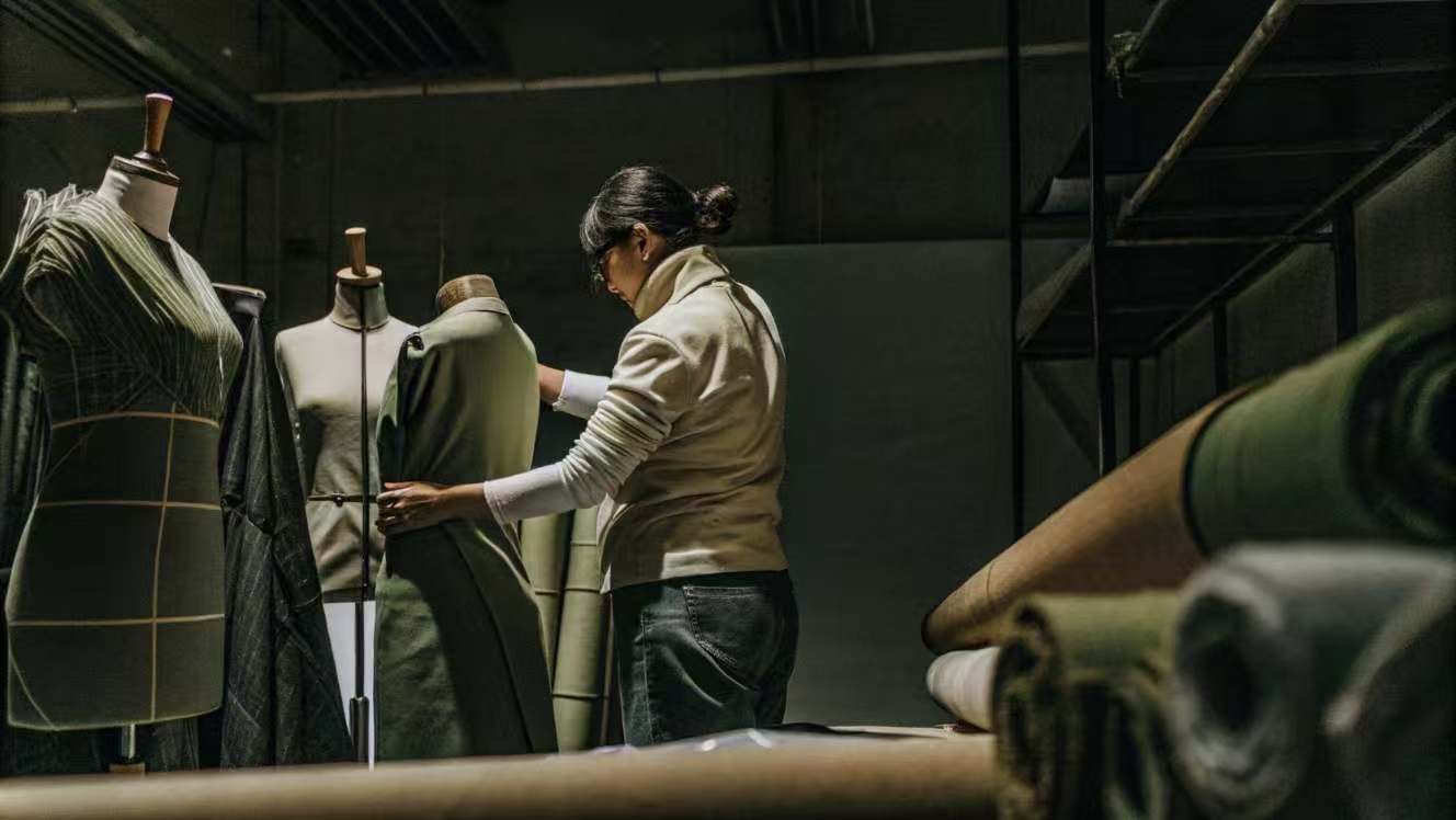 Designer fitting a green garment on a mannequin in a dimly lit workshop with fabric rolls.