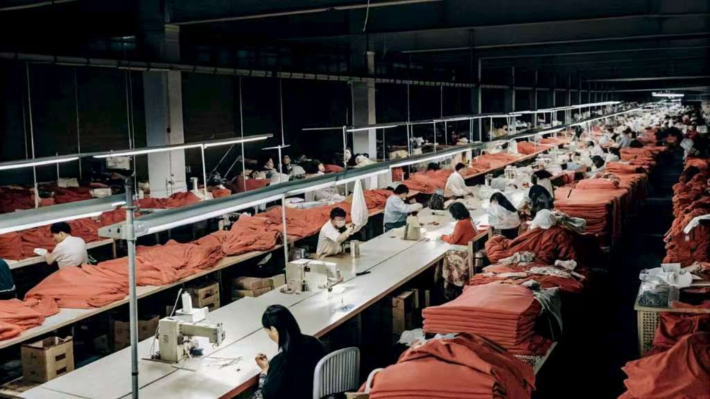 Factory workers sewing red fabric garments at long tables.