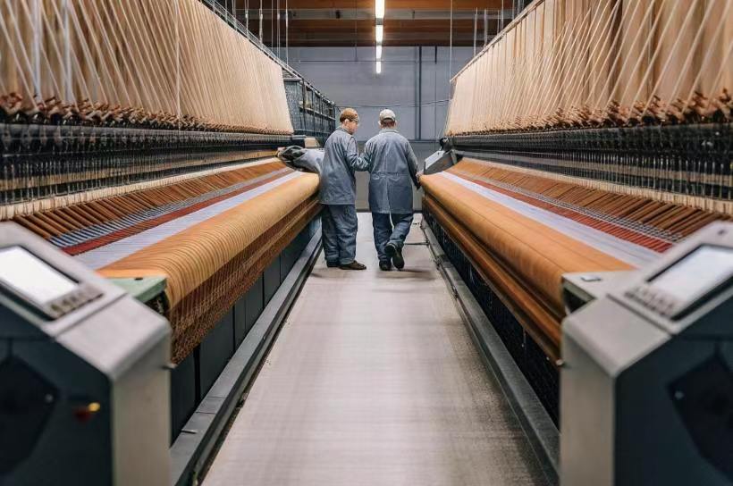 Two workers walk between large automated looms in a high-tech textile weaving facility.