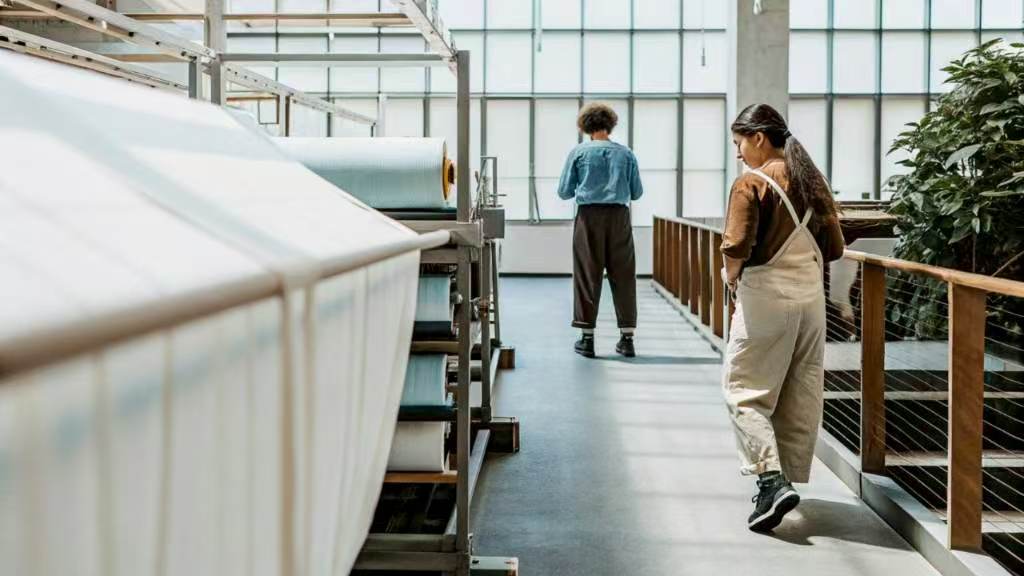 Two people walking through textile storage with fabric rolls on shelves
