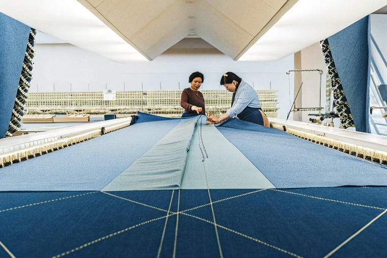 Two women collaborate on fabric manipulation in a textile workshop, working on a large blue fabric piece.